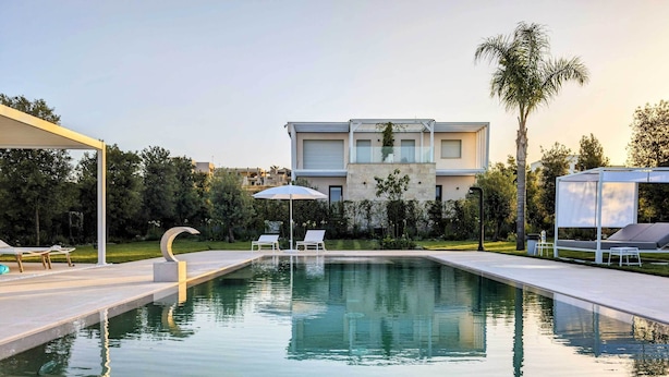 A modern pool area with lounge chairs and umbrellas, surrounded by a well-maintained lawn and a large building in the background. - Alternative Relax Rooms by Oikia Vacanze (Scorrano)