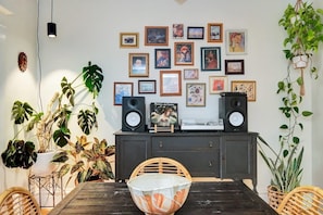 Uma sala de jantar com mesa de madeira, cadeiras, uma tigela grande, um armário escuro com quadros e diversas plantas em vasos.