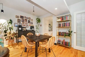 A dining area with a wooden table and wicker chairs, a bookshelf with books, and a kitchen area in the background.