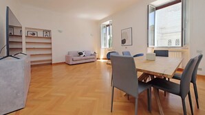 A spacious room with a wooden floor, a dining table with chairs, a sofa, and a large window with a view. - Luxury home in the Ghetto's heart (Roma)