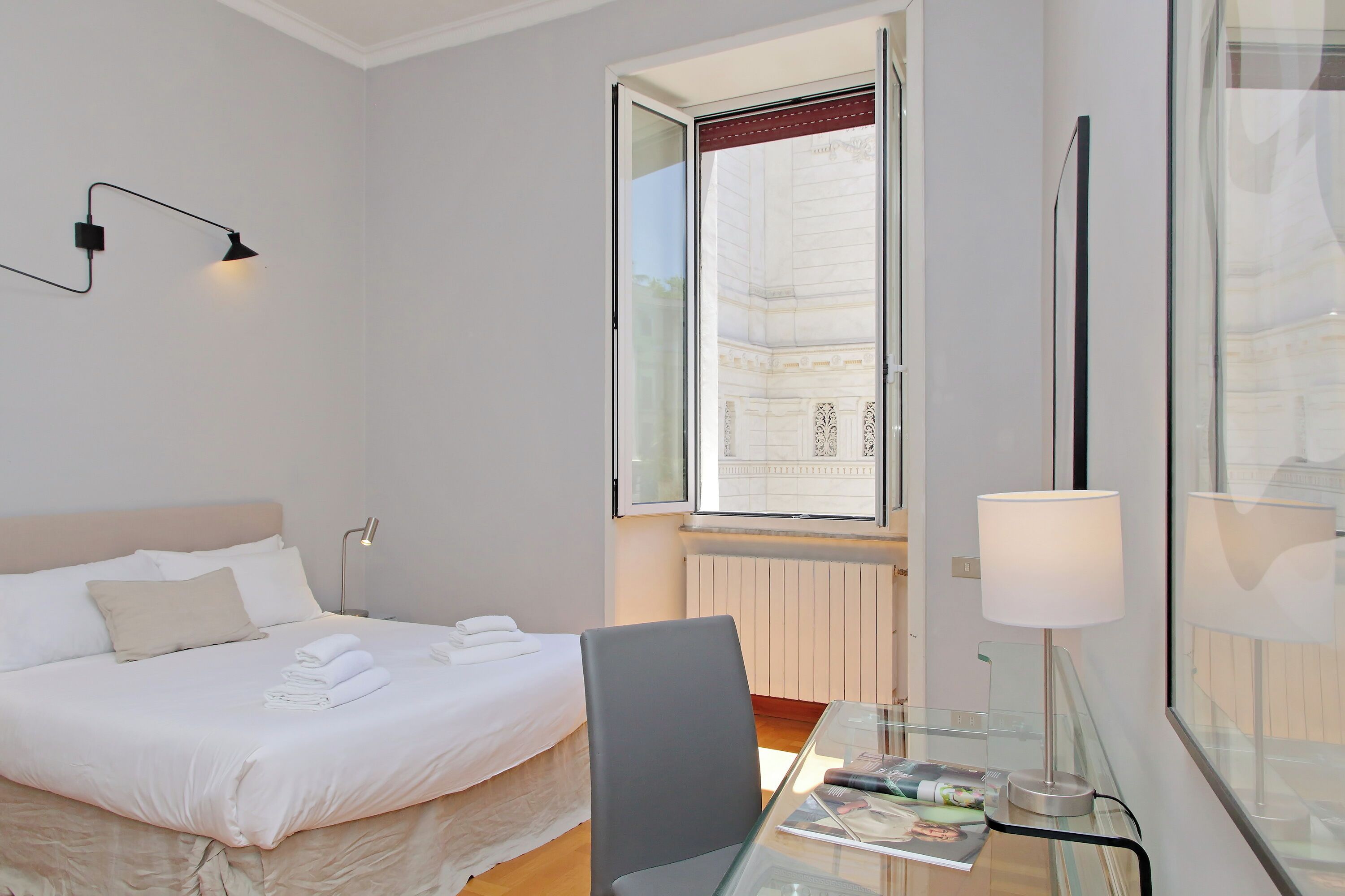 Une chambre d’hôtel avec un lit, une chaise, une table en verre, une lampe et une fenêtre donnant sur un immeuble.
