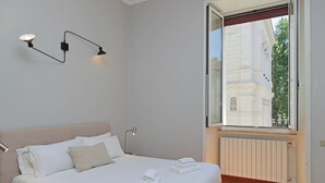 A bedroom with a bed, two wall-mounted lamps, a window with a view of a building, and a radiator. - Luxury home in the Ghetto's heart (Roma)