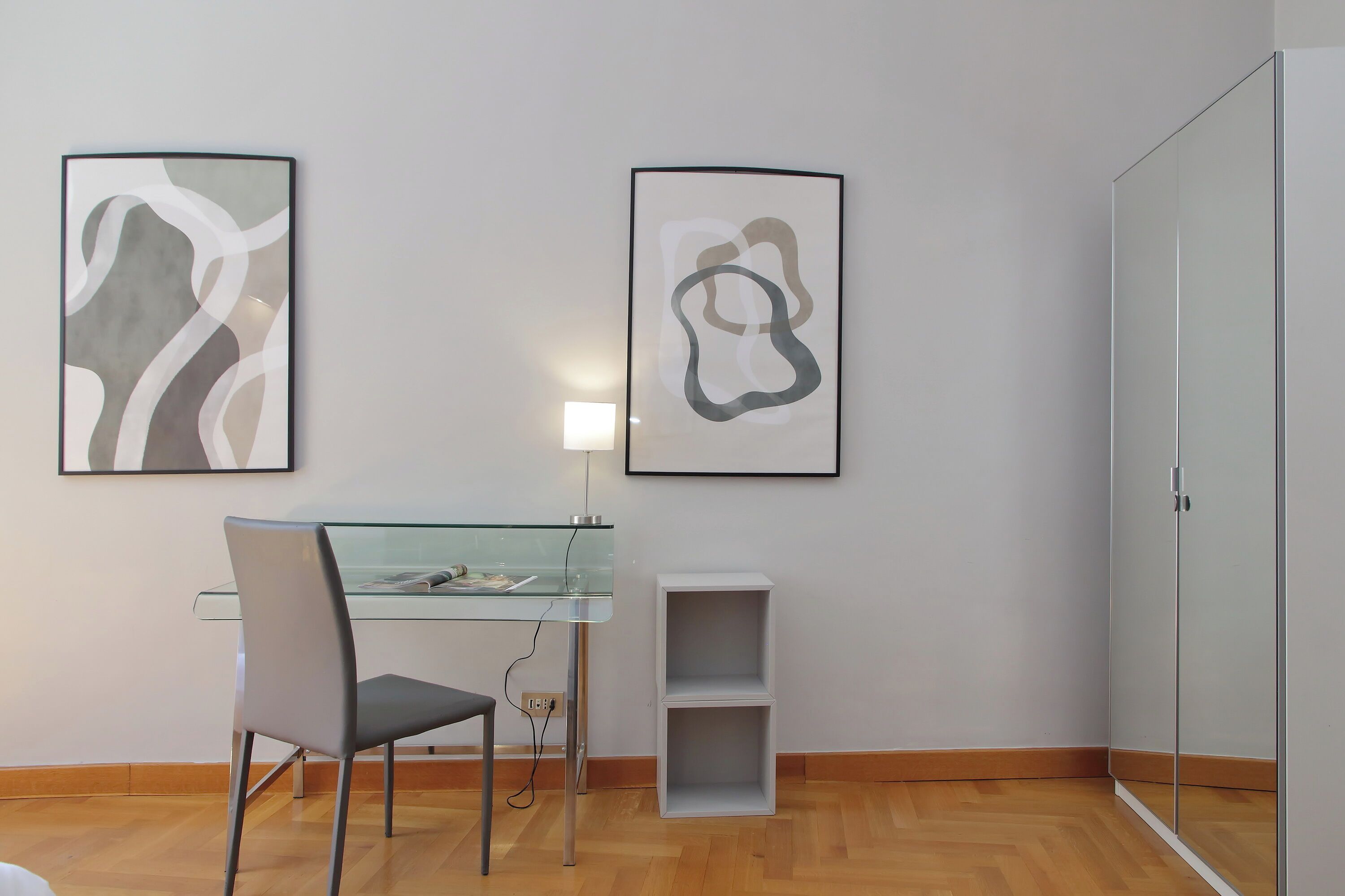 A modern room with a glass dining table, a grey chair, two abstract art pieces on the wall, a white cabinet, and a wardrobe.