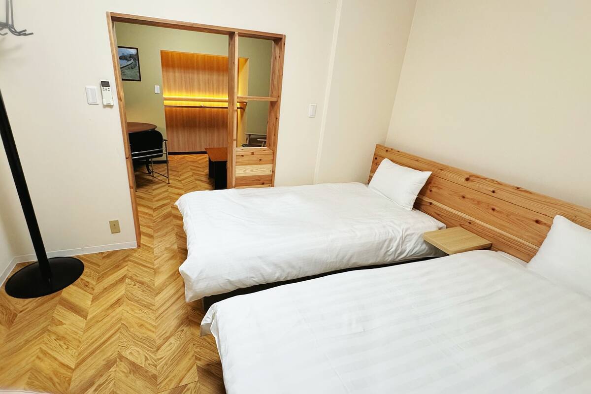 A hotel room with two beds, a wooden floor, a wardrobe, and a desk with a chair.