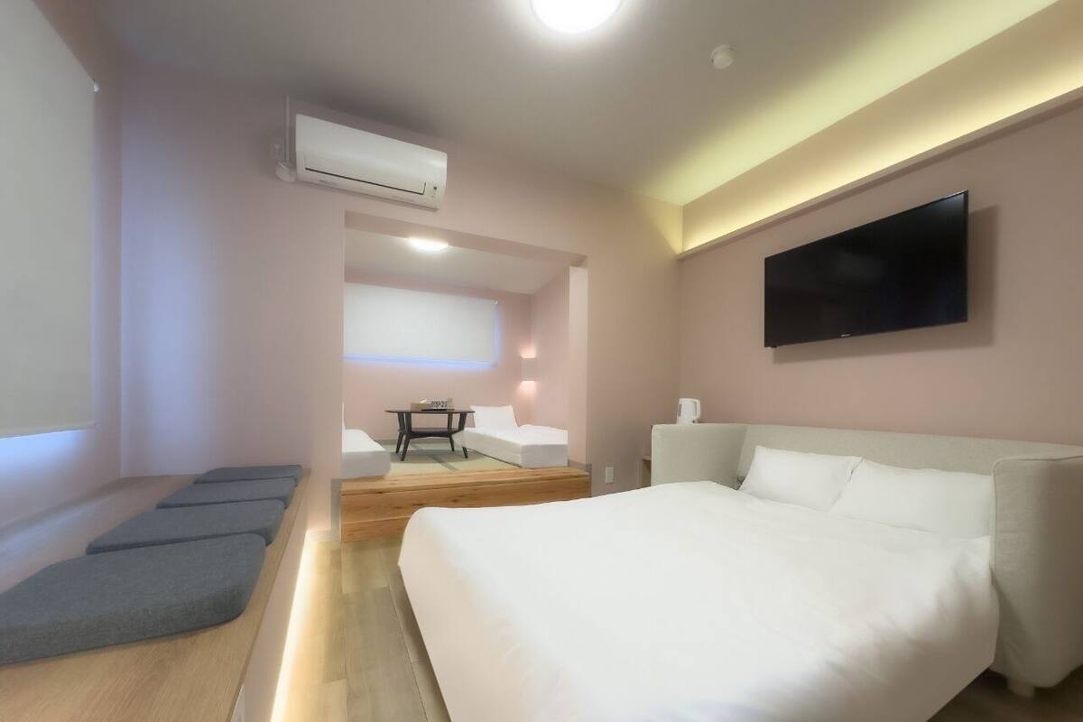 A modern hotel room with a large bed, a flat-screen TV mounted on the wall, a small table, and a seating area with cushions.