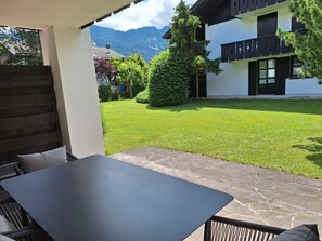 Un espace patio aménagé avec une table et des chaises, un chemin en pierre et une vue sur un bâtiment blanc aux volets noirs, avec une montagne en arrière-plan.