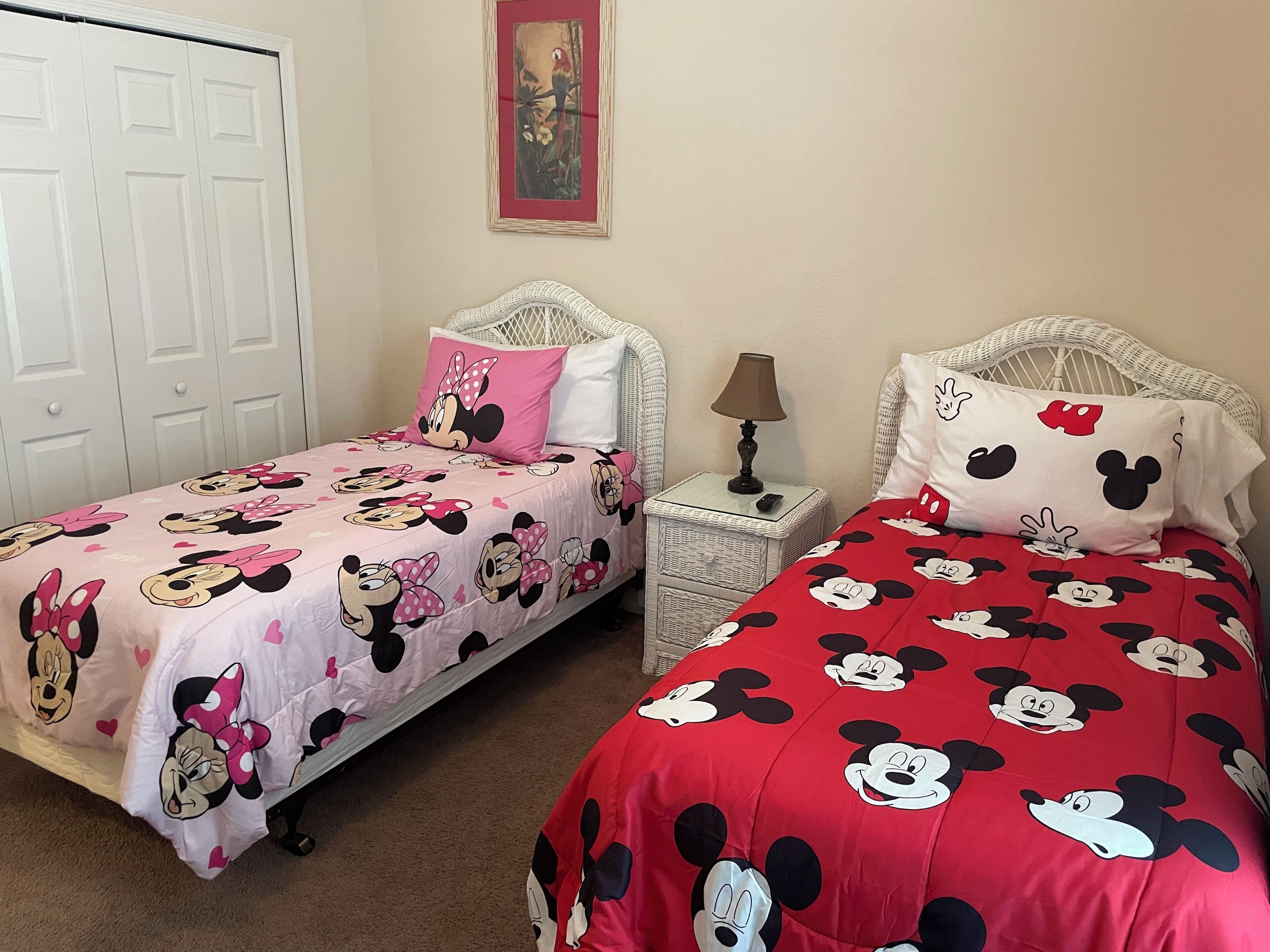 Deux lits jumeaux avec une literie aux motifs de Mickey Mouse, une porte blanche et une petite table avec une lampe.