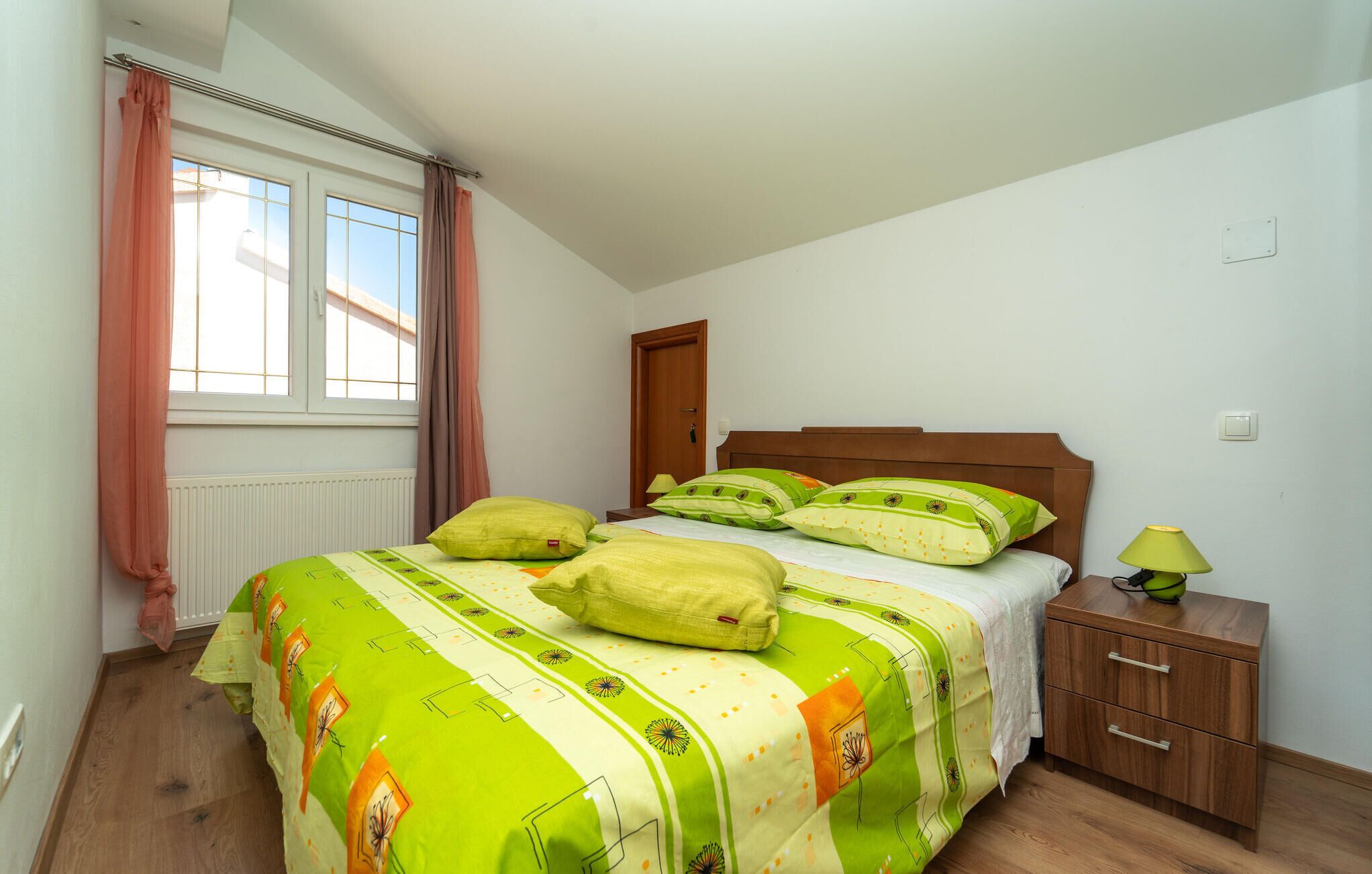 Een slaapkamer met een bed, een nachtkastje, een lamp, een raam met gordijnen en een deur.