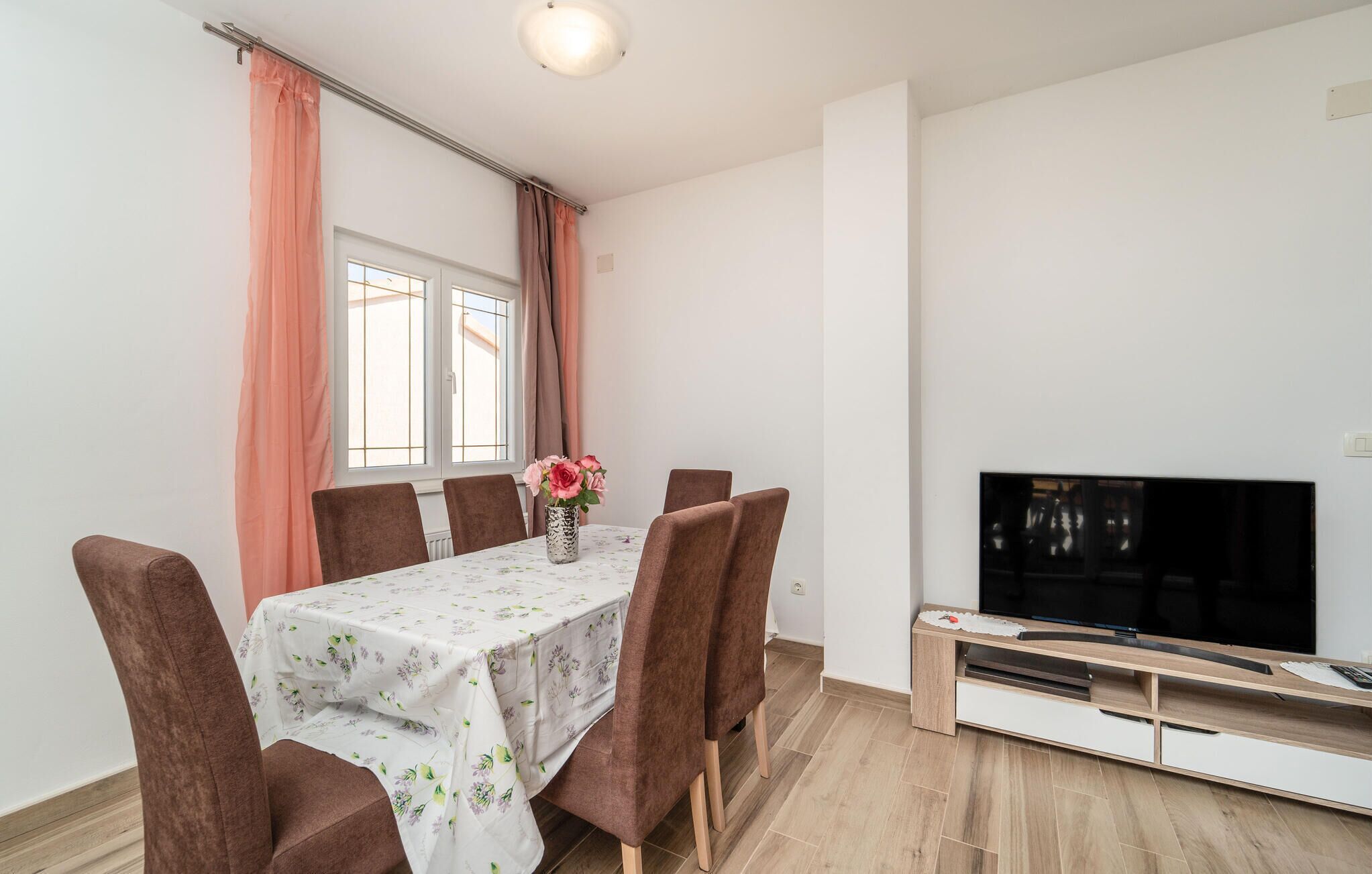 A dining area with a table set for two, a vase with flowers, and a TV on a wooden stand.