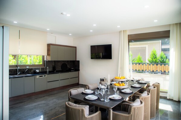 A modern kitchen with a dining area, featuring a large table set for six, a flat-screen TV, and a view of greenery outside. - Living Dream Villa Relax (Belek)