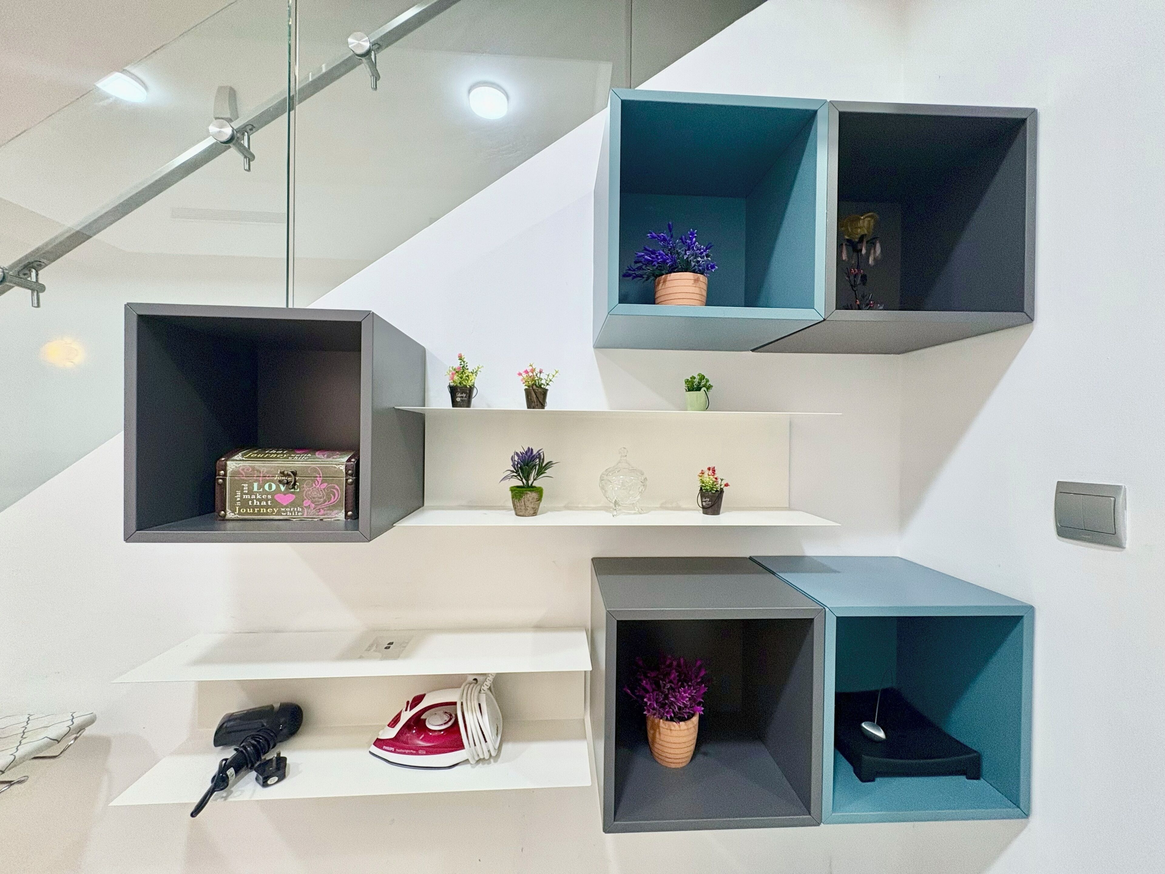 A modern staircase with built-in storage shelves holding various items like a suitcase, an iron, and decorative plants.