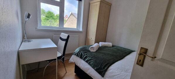 A small bedroom with a bed, a desk, a chair, a radiator, and a window with a view of houses. - Alexandra Palace 3 Bedrooms Apartment in London (London)