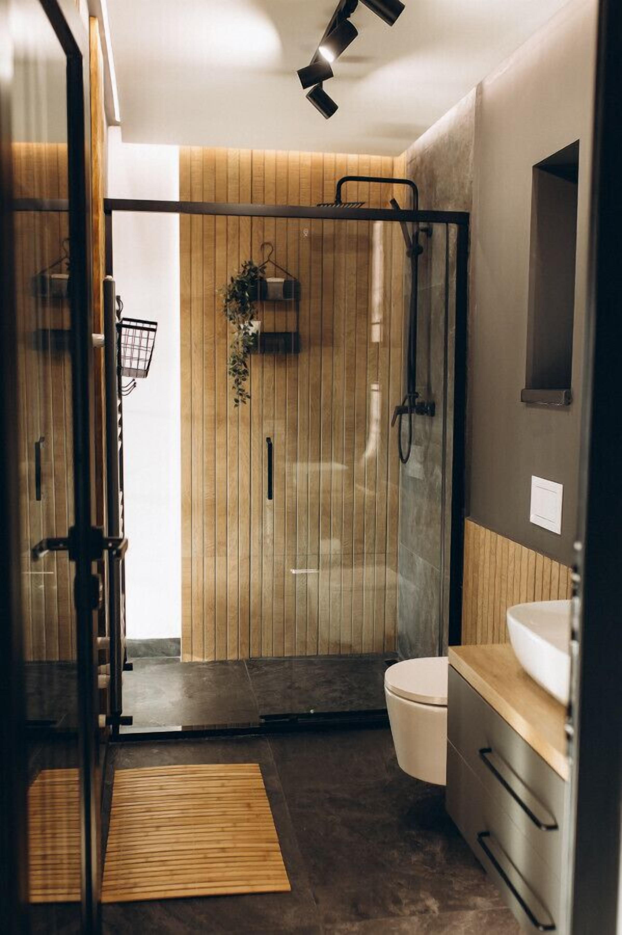 A modern bathroom with a glass shower enclosure, wooden panel walls, and a wooden floor mat.