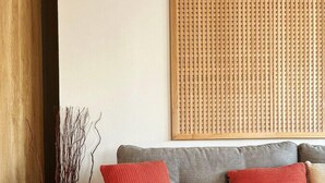 Ein graues Sofa mit zwei roten und einem beigen Kissen, darüber ein gewebtes Wandbild und ein Holzboden.