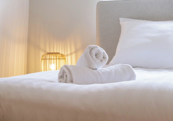 A neatly made bed with a folded white towel on top, a bedside lamp, and a white pillow.