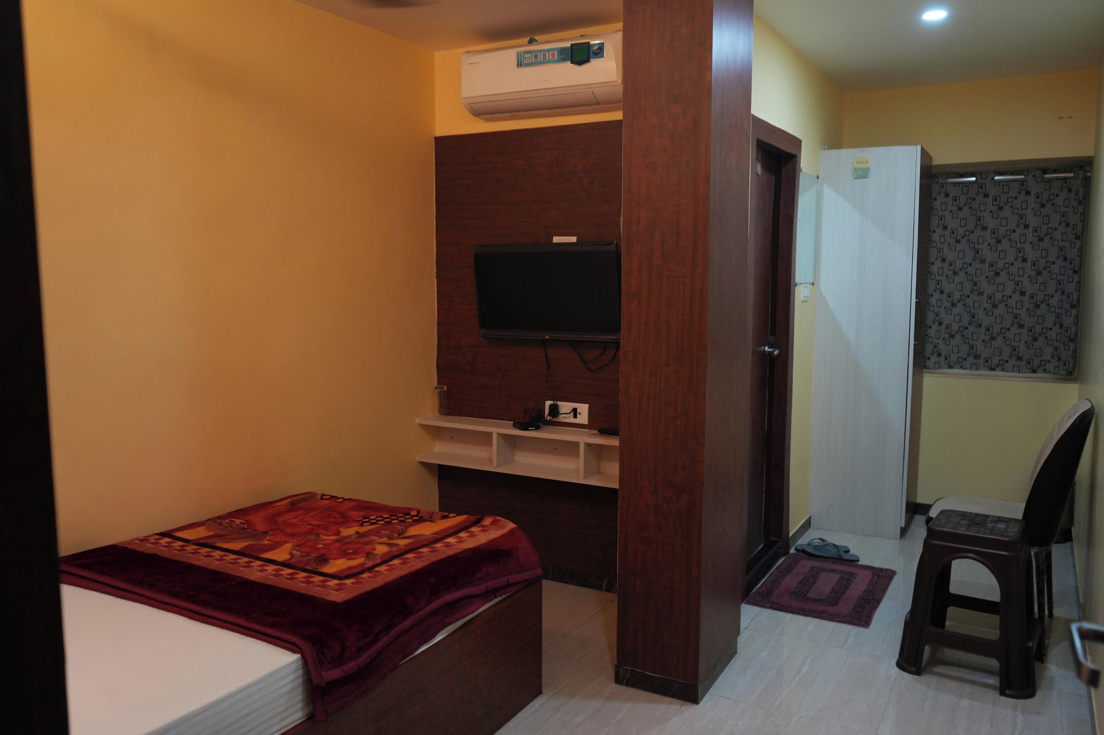 Una habitación pequeña con una cama, un televisor fijado a un panel de madera, un refrigerador blanco y una silla.