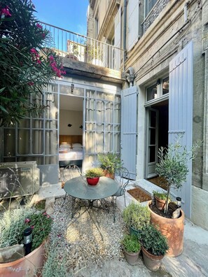 A courtyard with a round table, potted plants, and a building with grey shutters. - Le Jardin des Etudes - Terrace & Mansion 300 Years (Avignon)