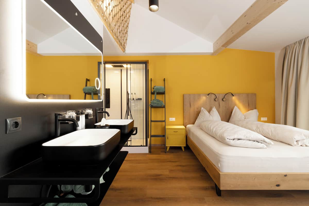 A modern bedroom with a large bed, a bedside table, a nightstand, a mirror, and a bathroom with a sink and towel rack.