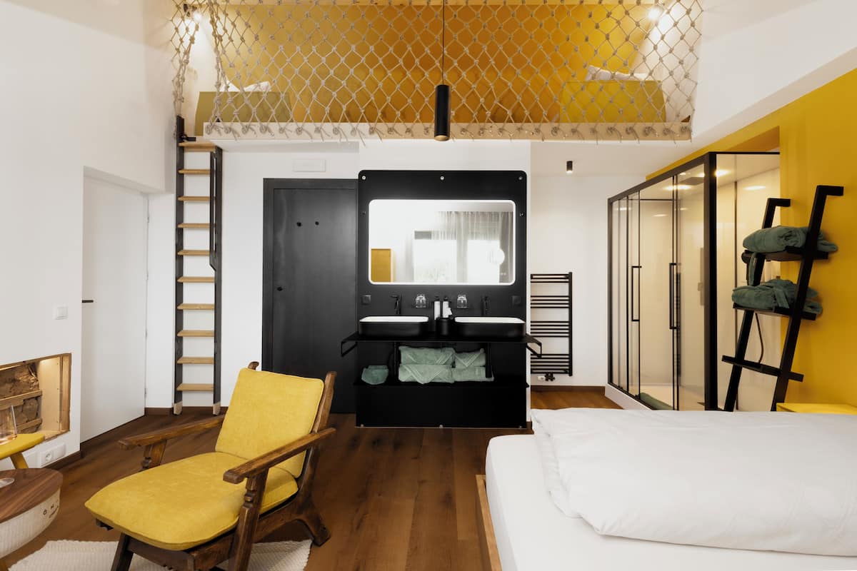 A compact bedroom with a bed, a yellow chair, a wooden ladder, and a black wardrobe with a mirror.