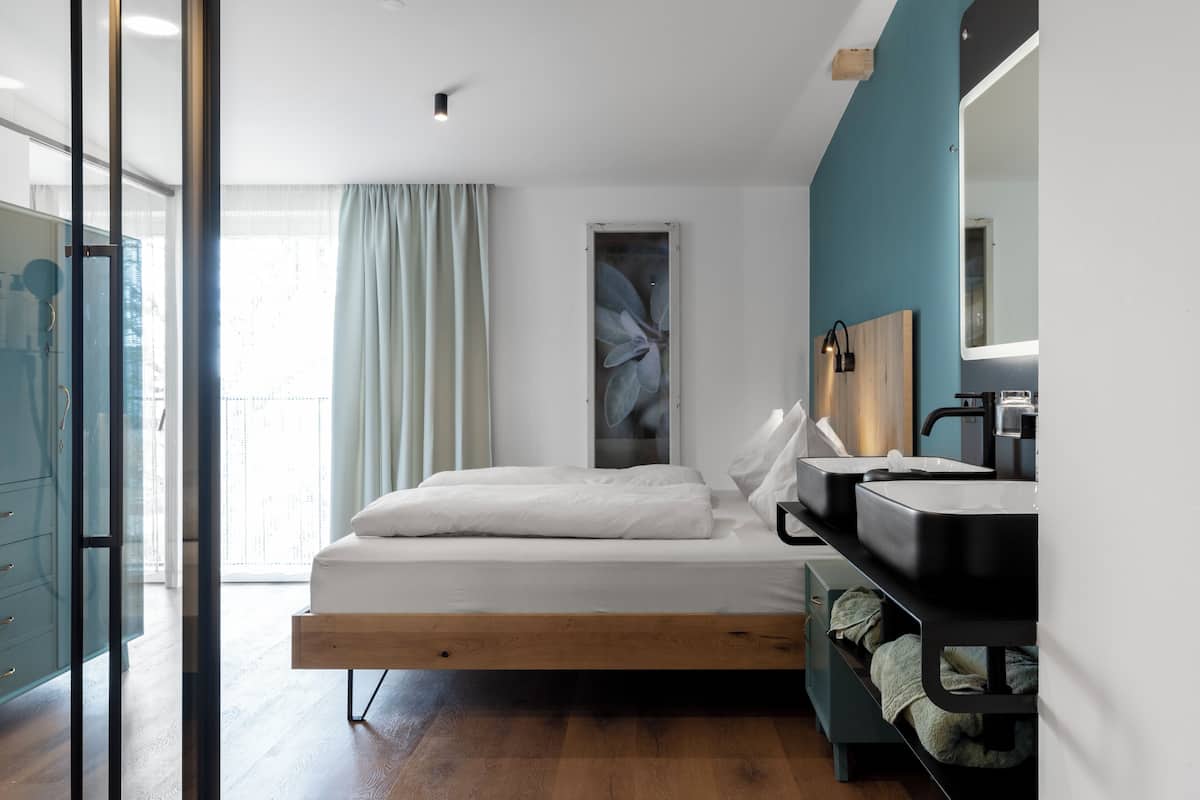 A modern bedroom with a bed, bedside tables, a built-in bench, a large window with curtains, and a bathroom with a mirror and sink.