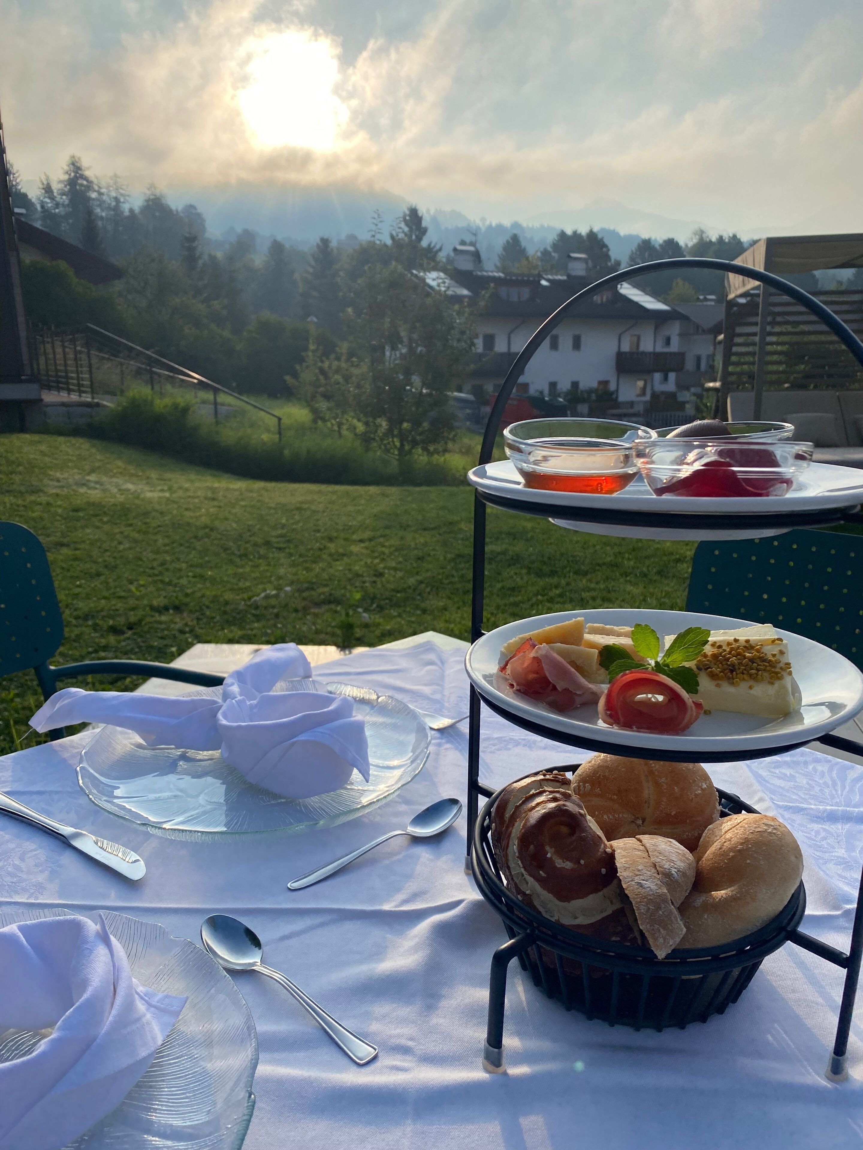 Mesa de comedor al aire libre con tres niveles, platos de comida, una cesta de pan y una vista panorámica de una casa y árboles al fondo.
