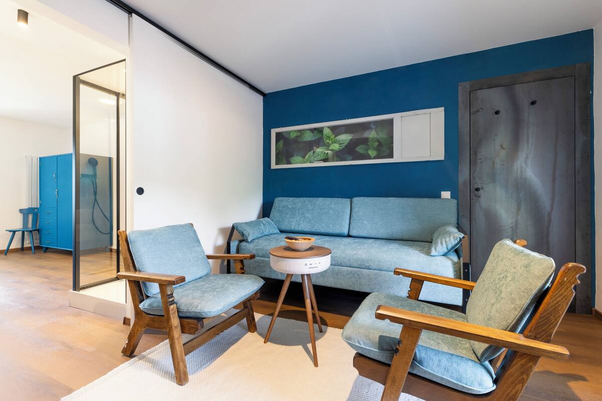 A modern living room with a blue sofa, two wooden armchairs, a small round table, and a glass door leading to another room.