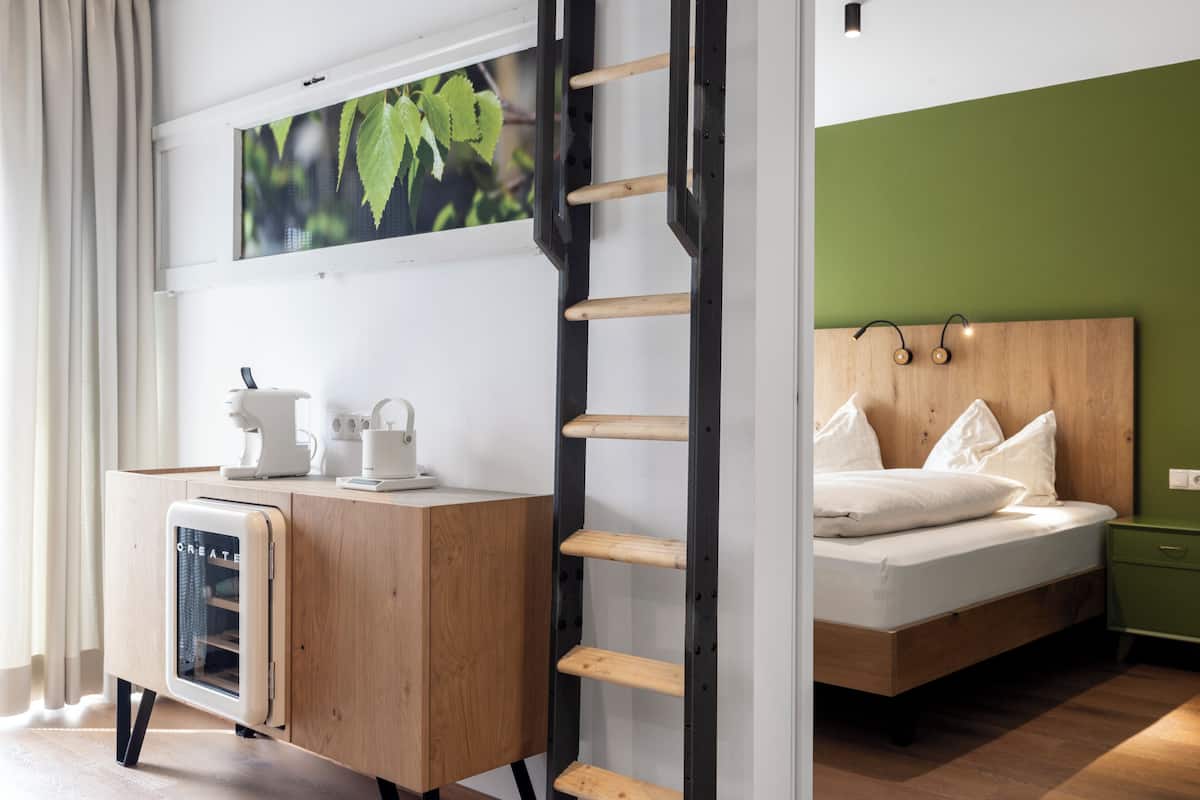 A modern hotel room with a wooden desk, a bed with white bedding, a green accent wall, and a window with a view of greenery.