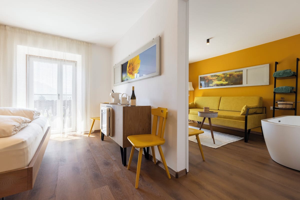 A modern hotel room with a bed, a yellow sofa, a white armchair, and a dining area with a table and chairs.