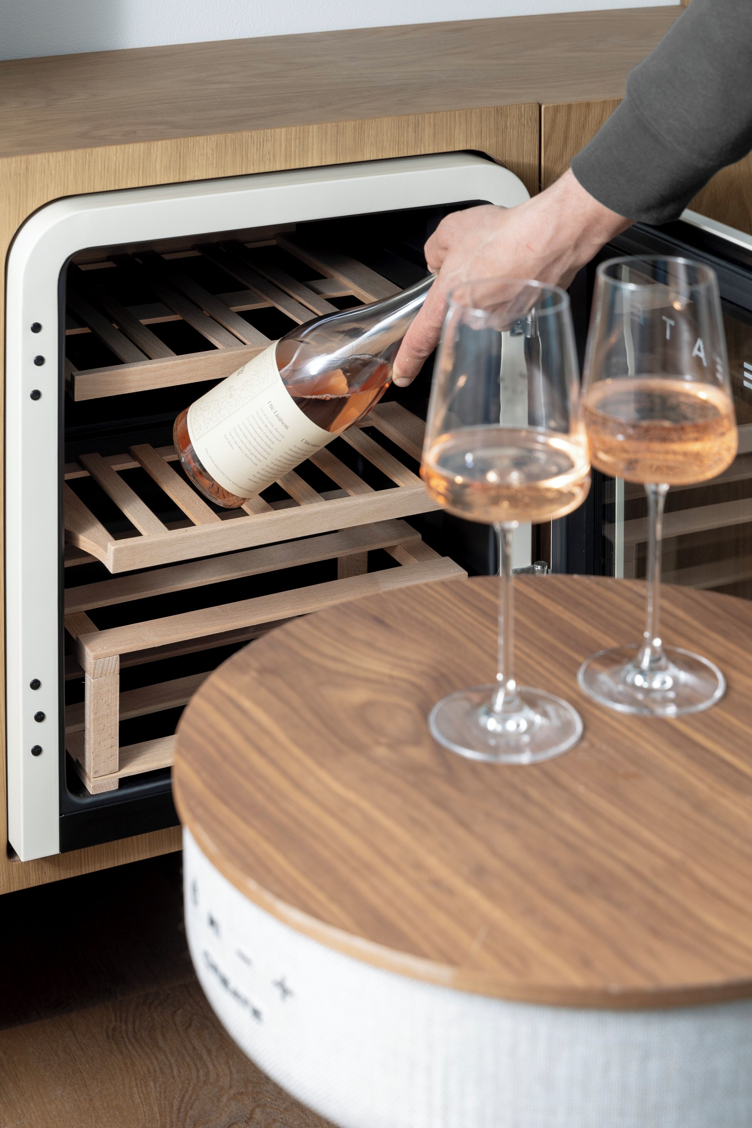 A wine bottle being poured into a wine cooler with two glasses on a wooden table.