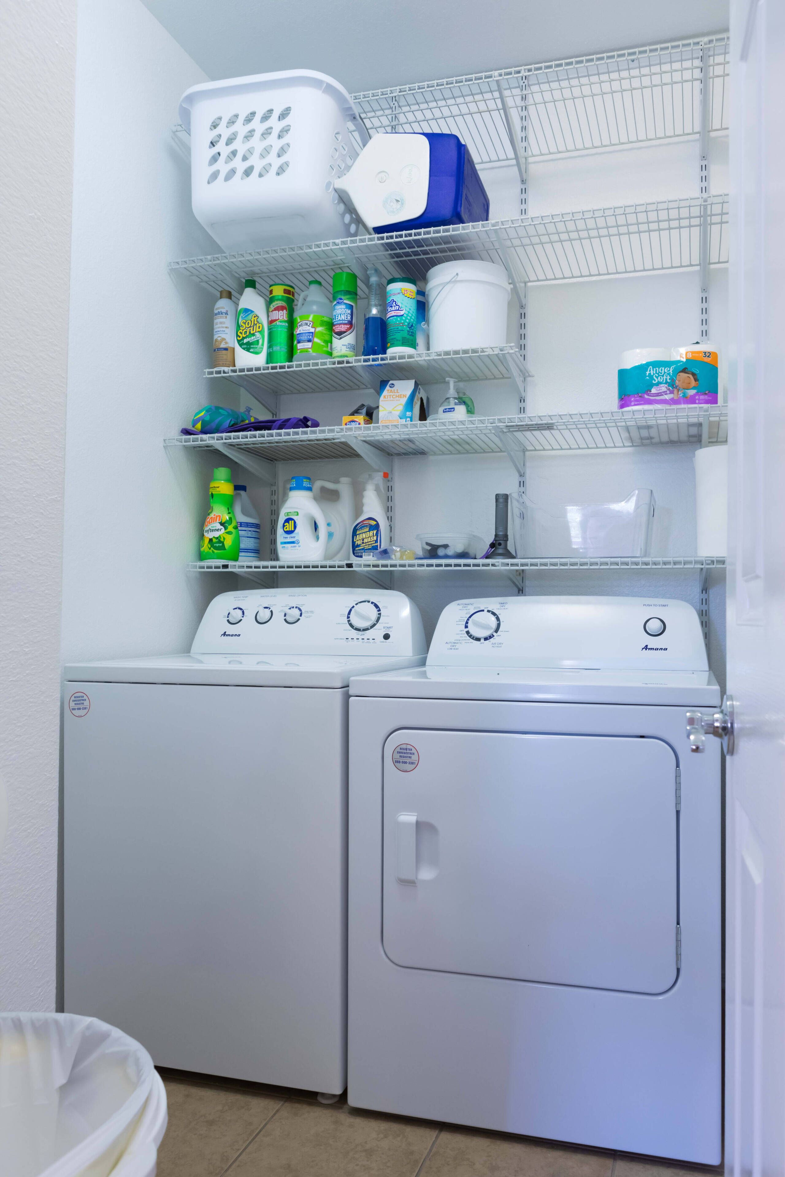 A laundry area with two front-loading washing machines, shelves with various cleaning products, and a basket.