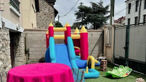 A backyard with a small inflatable slide, a round table with a pink tablecloth, wicker chairs, and a green play area. - Quiet house between Disneyland and Paris (Villiers-sur-Marne)