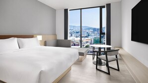 A modern hotel room with a large bed, a desk, and a view of a cityscape through large windows.