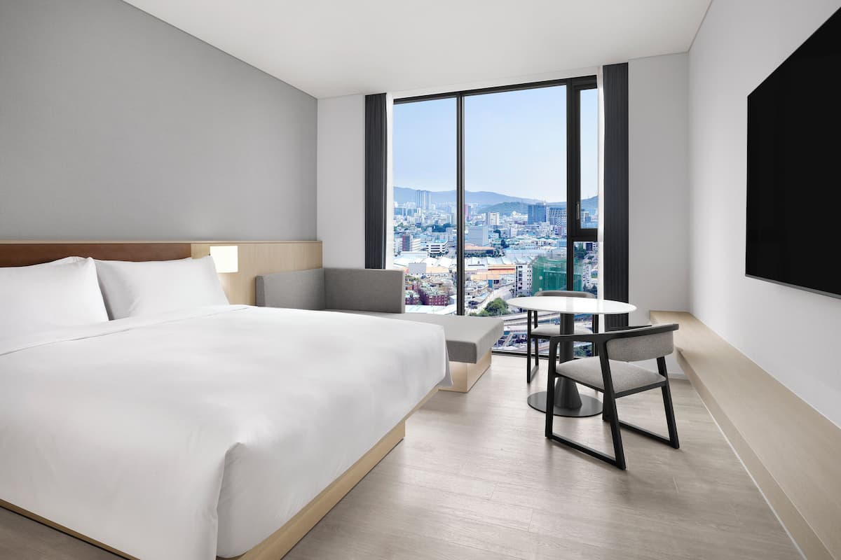 A modern hotel room with a large bed, a desk, and a view of a cityscape through large windows.