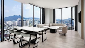 A modern living room with a large table and chairs, a sofa, and a view of a cityscape through large windows.