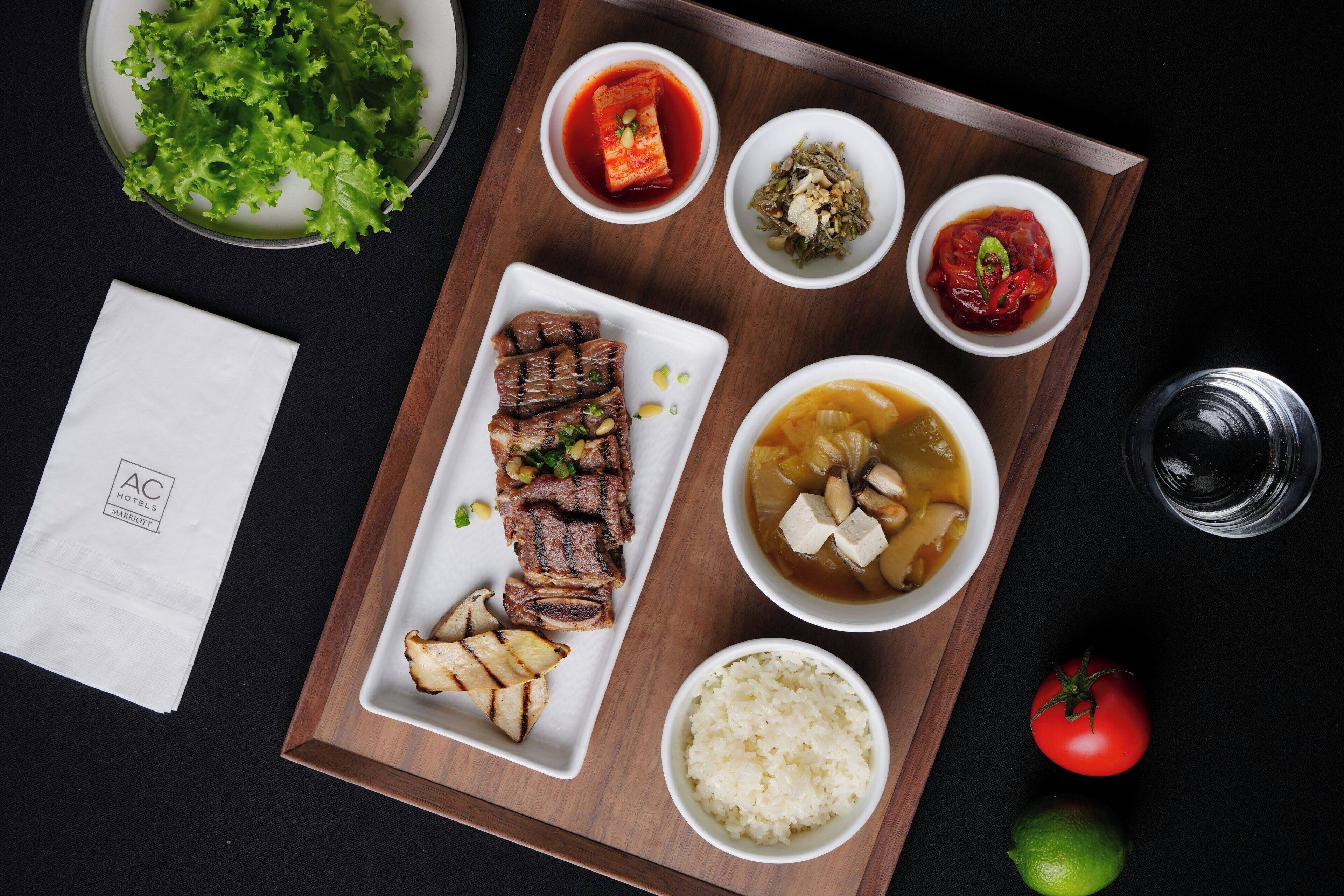 A tray with grilled meat, rice, and various side dishes, including kimchi and a soup with tofu.