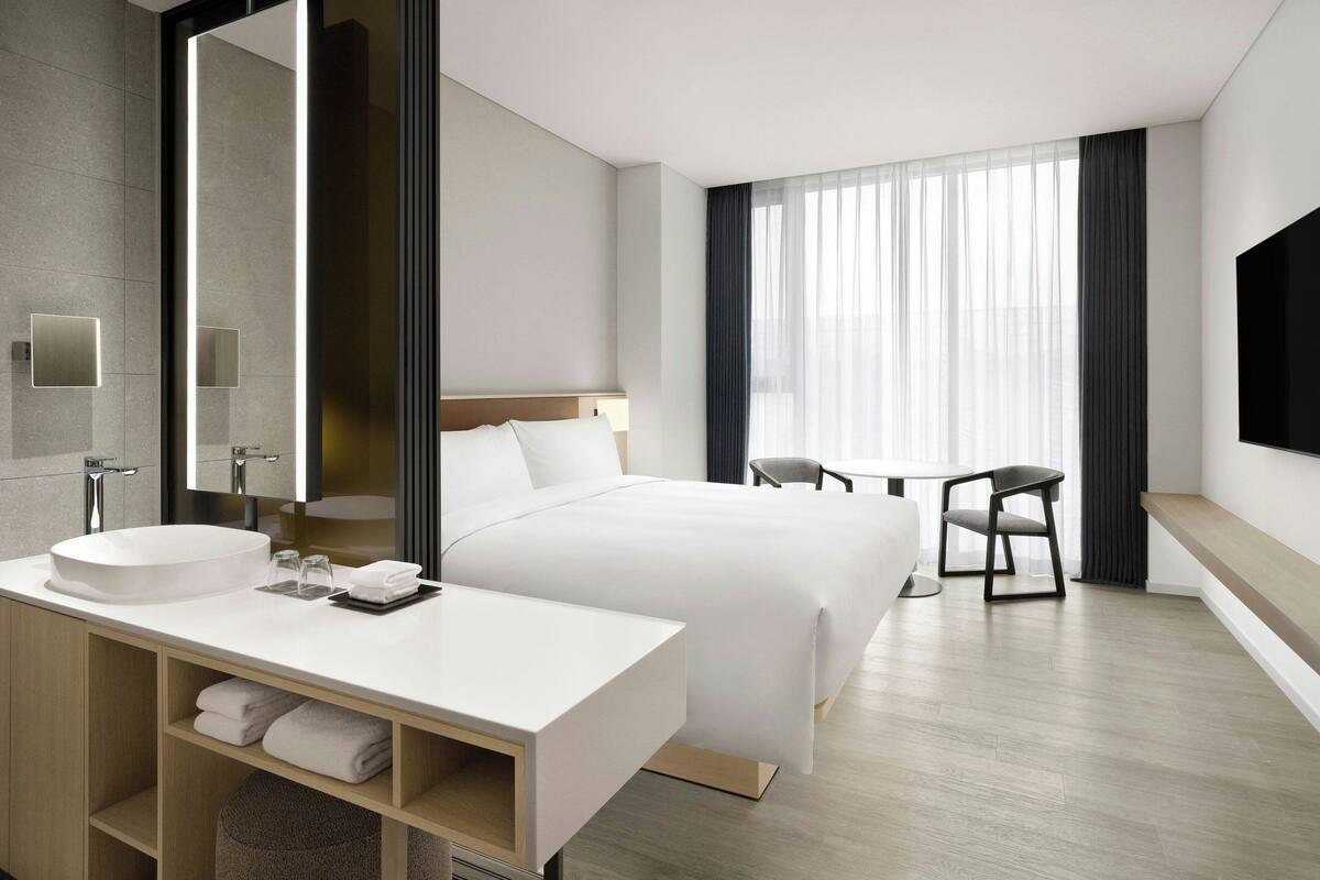 A modern hotel room with a large bed, a bedside table with a white sink, a mirror, a flat-screen TV, and a window with curtains.