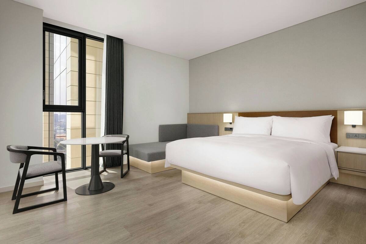 A modern hotel room with a large bed, a small table, two chairs, a bench, and a large window with curtains.