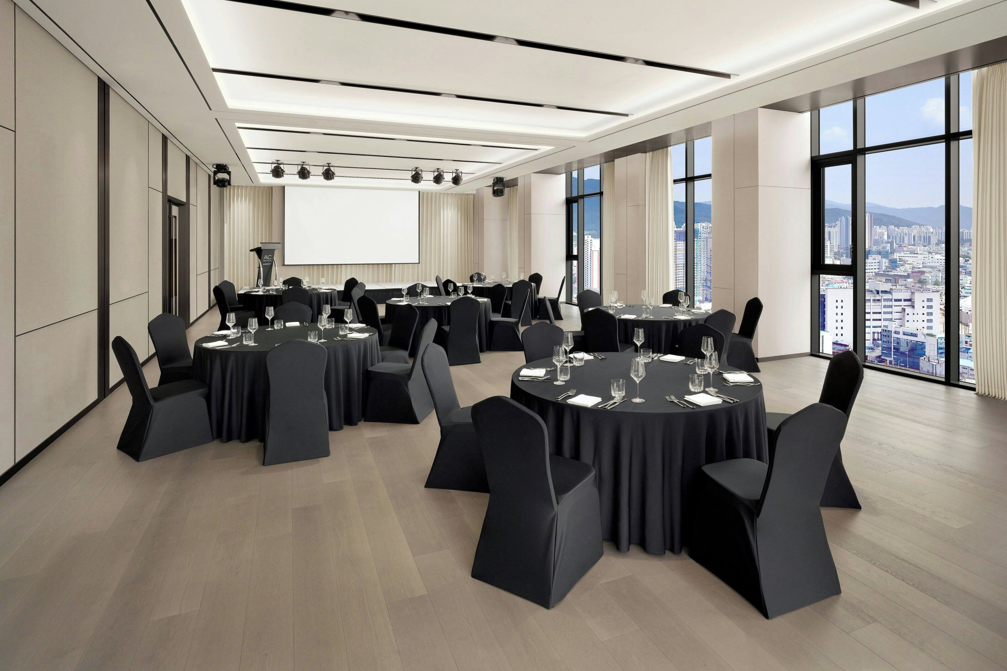 A conference room with multiple tables, black chairs, and a large screen.