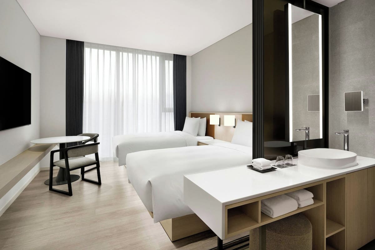 A modern hotel room with two beds, a round white sink, a large mirror, a flat-screen TV, and a dining table with chairs.