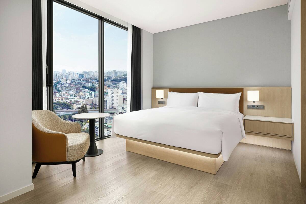 A modern hotel room with a large bed, a small round table, a chair, and a view of the city through large windows.