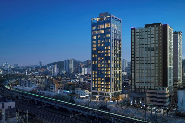 Un hôtel moderne à plusieurs étages, de grande hauteur, entouré d’un paysage urbain et bordé par une voie ferrée.