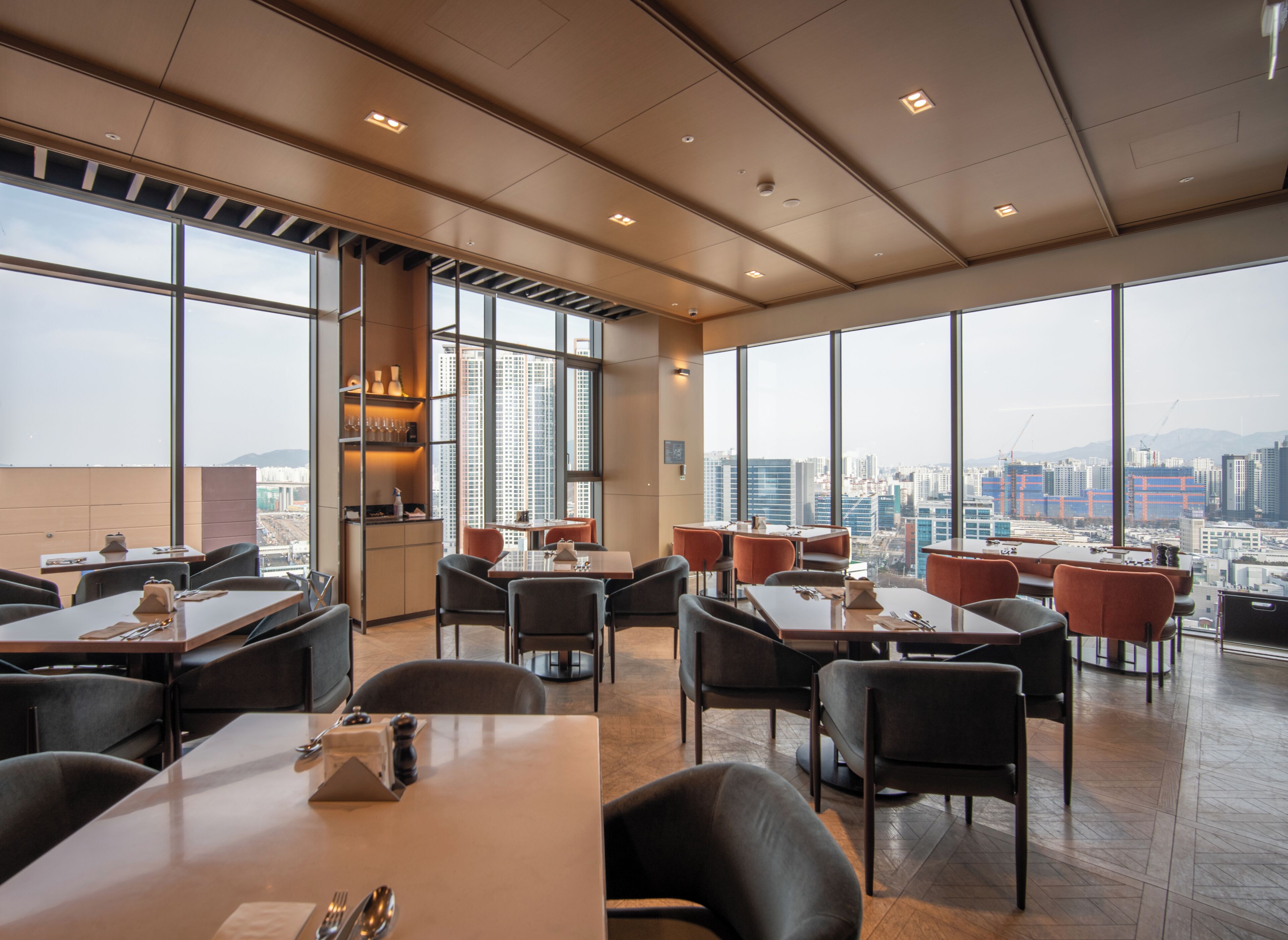A modern dining area with tables and chairs, large windows offering a city view, and a high ceiling with recessed lighting.