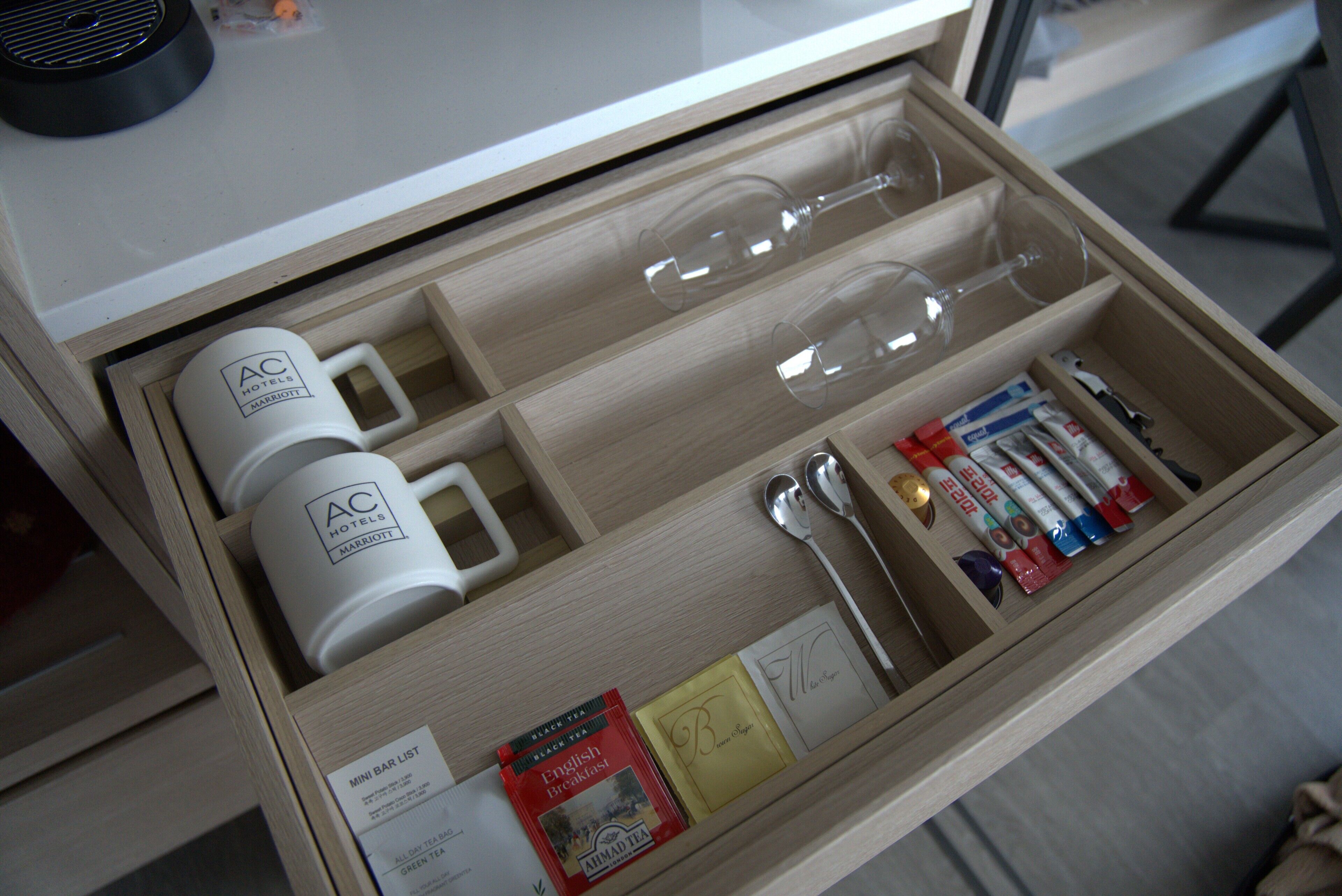 A hotel minibar drawer with various items including glasses, a mini bar list, tea packets, and a spoon.