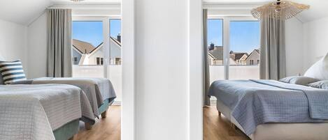 A modern bedroom with two beds, large windows, and a view of houses.