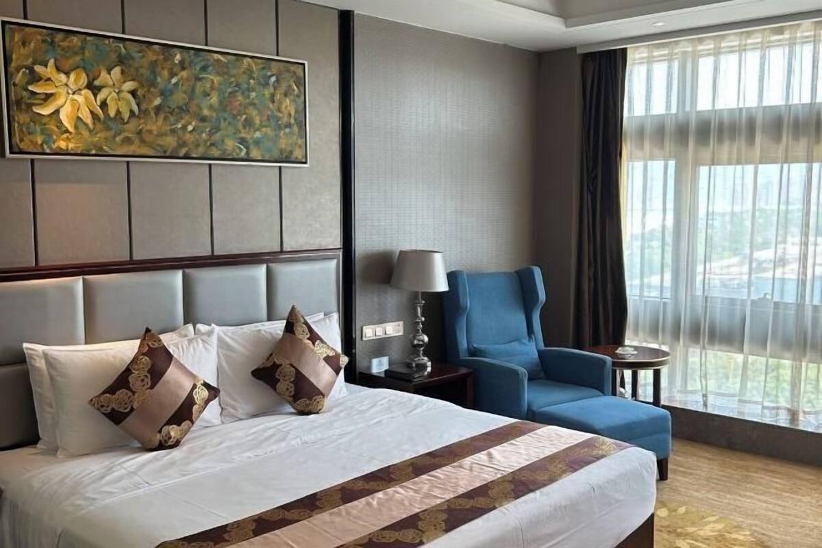A hotel room with a large bed, a blue armchair, a painting, and a view through the window.