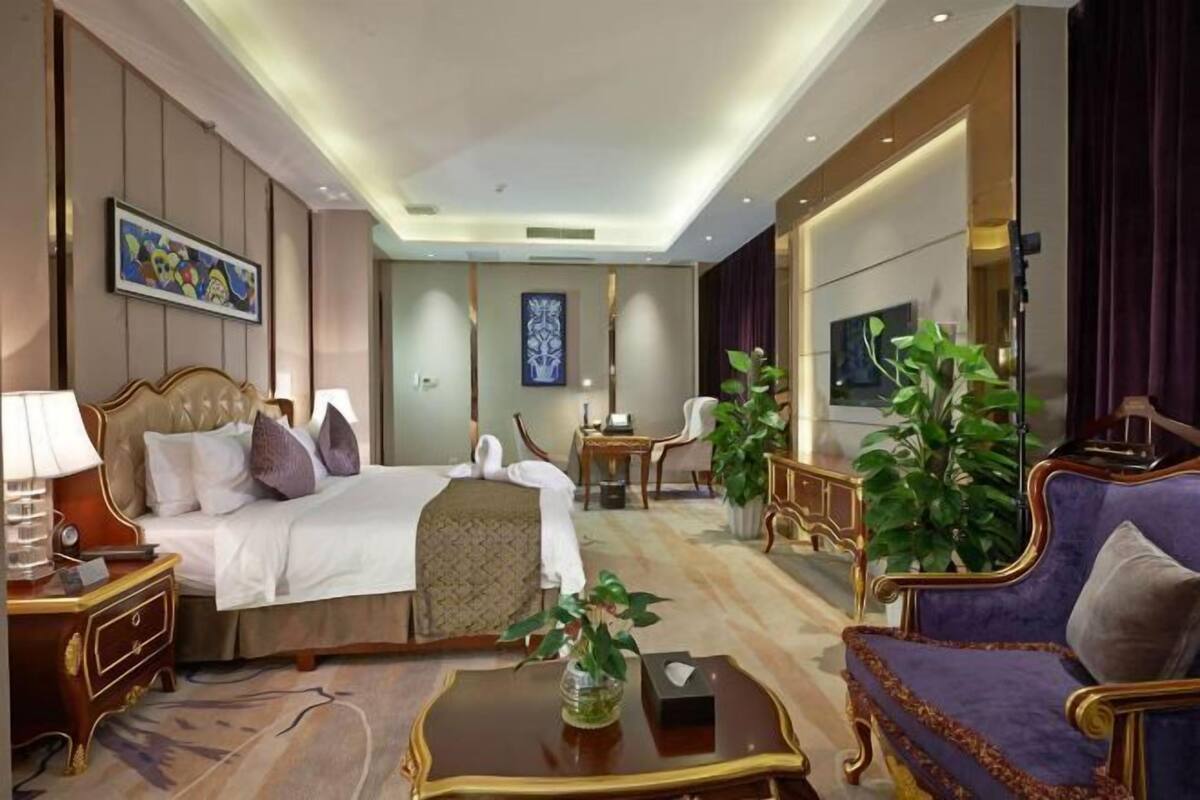 A hotel room with a bed, a sitting area with a sofa and armchairs, a coffee table, a TV, and a large window.