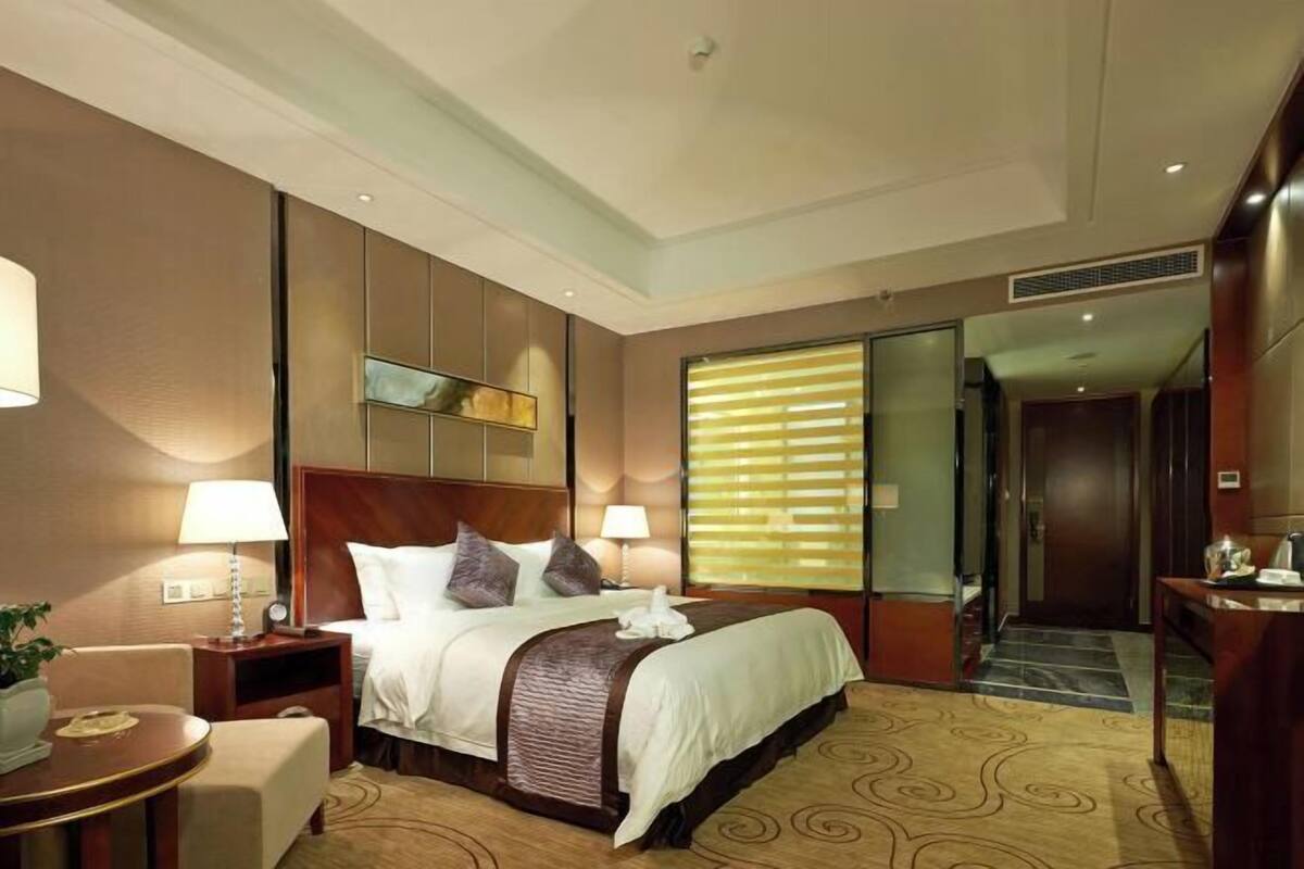 A hotel room with a bed, bedside tables, a desk, a chair, a lamp, a potted plant, and a window with blinds.