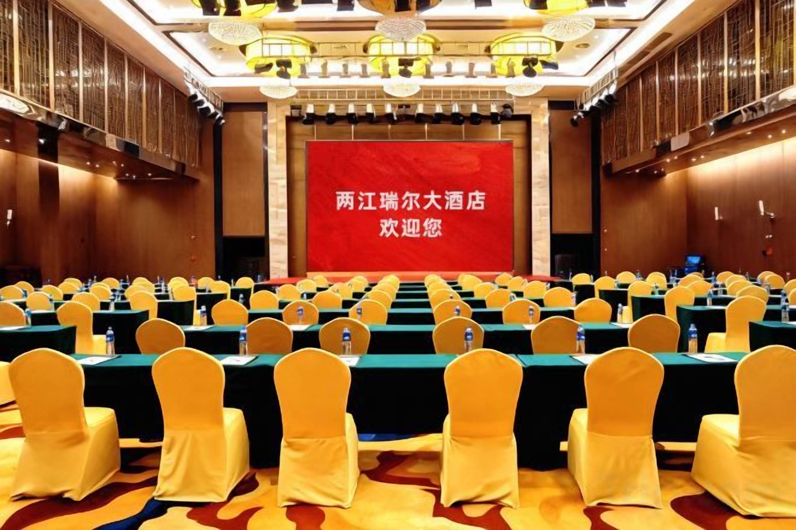Un salón de conferencias con filas de mesas y sillas, una pantalla grande y un banderín rojo.