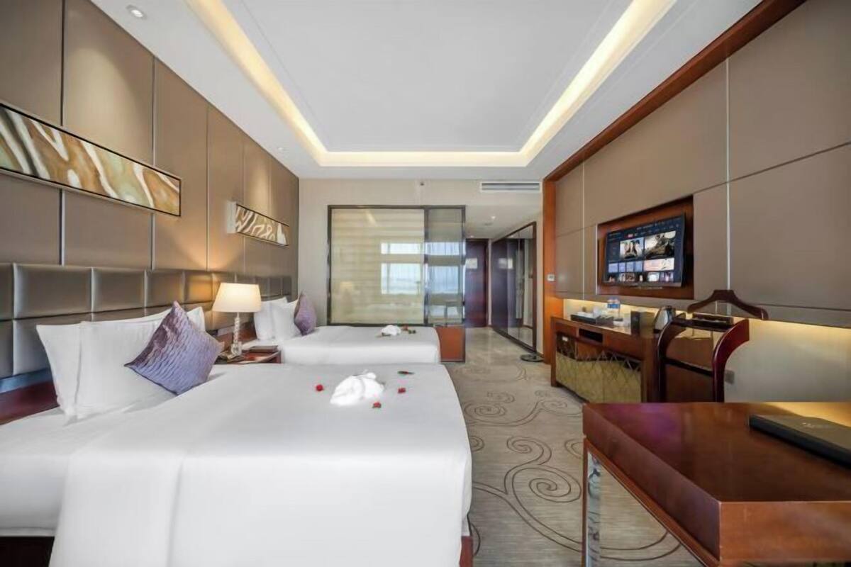A hotel room with two beds, a desk, a television, and a view of the city.
