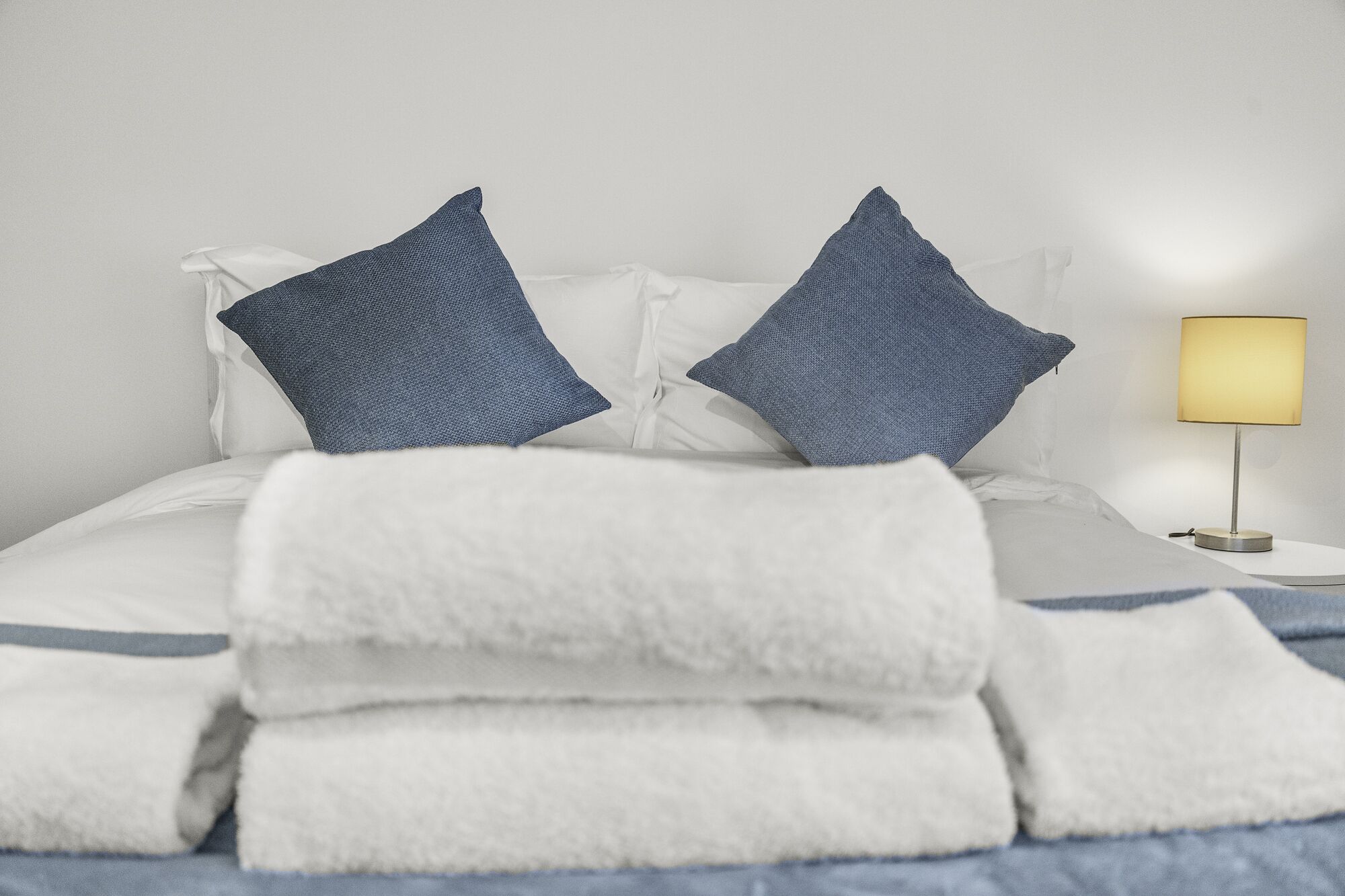 Un lit bien fait, avec du linge de lit blanc et deux oreillers bleus, une lampe de chevet et un mur blanc.
